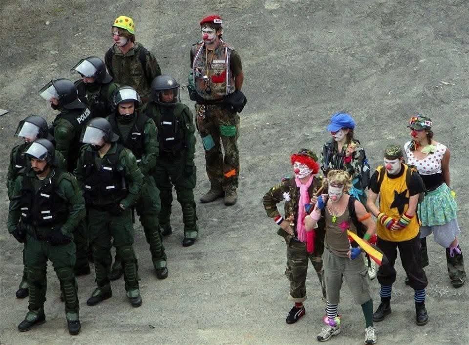 A military platoon standing next to a clown platoon. Two clowns have joined the military platoon, standing at the back, imitating the platoon’s tough stance