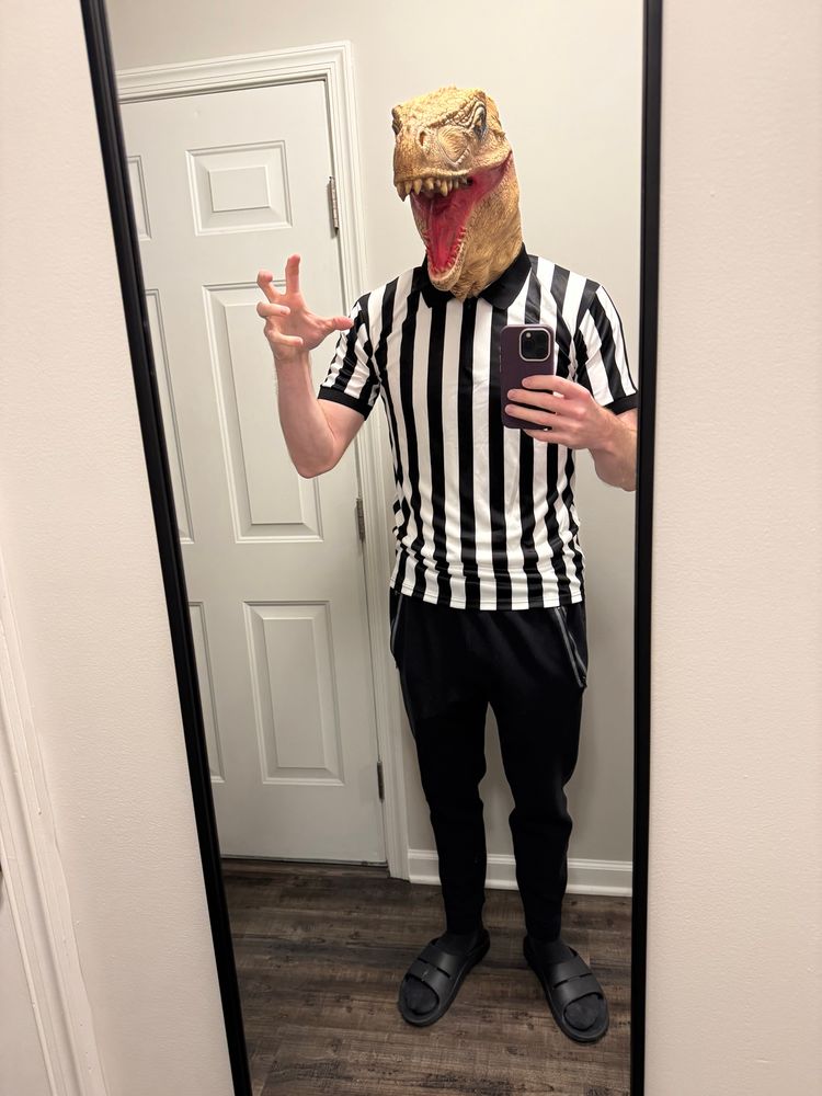 A white man stands in a mirror, wearing the black/white striped shirt and black pants of a football referee uniform. He wears a brown and tan roaring Tyranosaurus dinosaur mask. His right hand is up like a swiping claw.