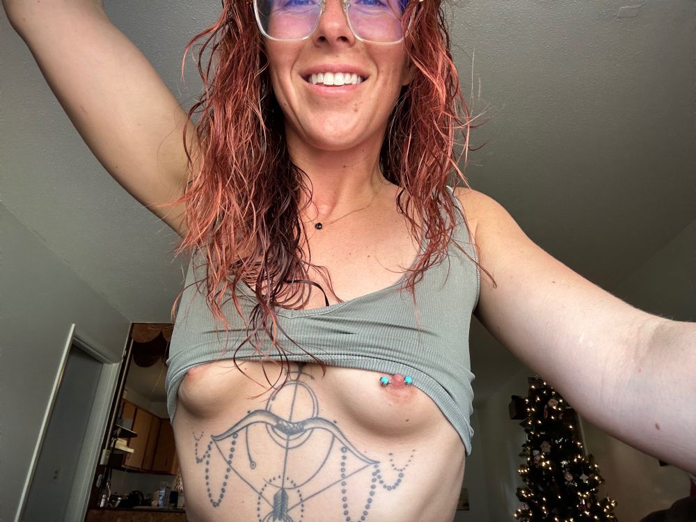 Red haired person (Summer!) with white teeth, small tits with a blue piercing, and tattoos on their torso