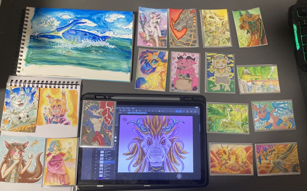 A picture of multiple pieces of art that were posted on my bluesky account during the month of July, most of them in trading card sized. There is also a tablet with a digital portrait of a dragon.