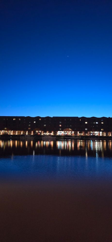Liverpool docks at dusk.  The sky is cloudless shading from light blue to almost black at the top. Jupiter visible in the sky.  Lights reflect off the water.
