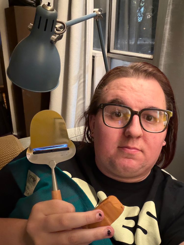 liz, lying on the couch under a blanket. she’s holding a cheese slicer and the remnants of a small block of brunost, a norwegian cheese made by caramelising the sugars in the milk used to produce it