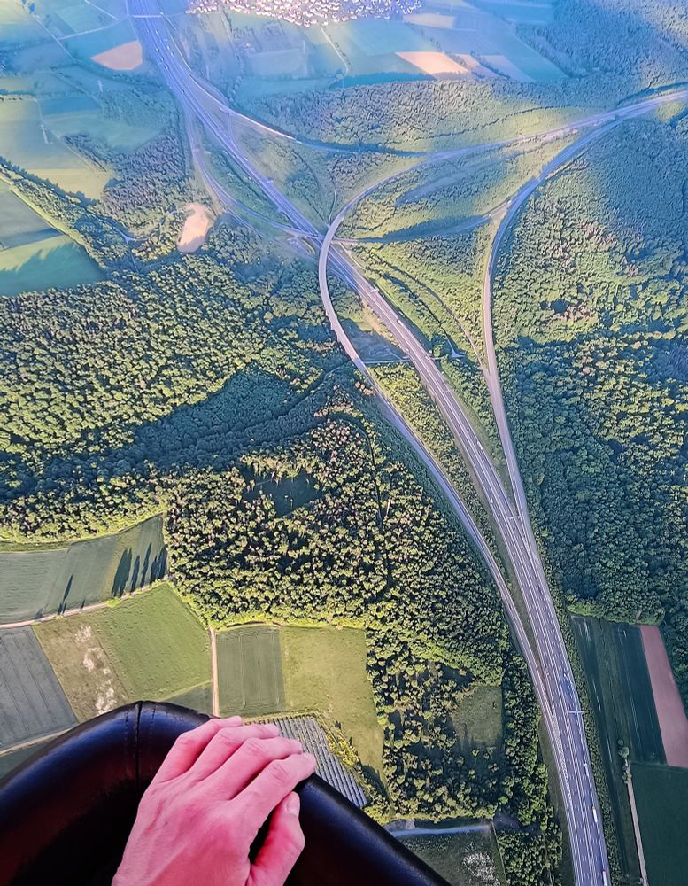 Blick aus einem Fesselballon auf die darunterliegende Landschaft mit Bäumen und einer Autobahn.
Auf dem Korbrand des Ballons ist eine Hand zu sehen.