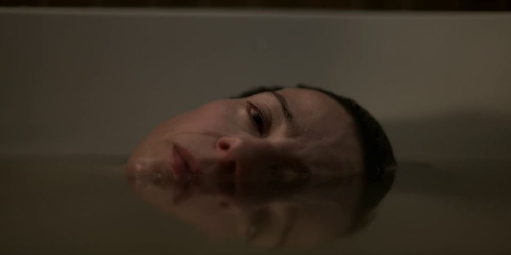 O rosto de Laura parcialmente coberto pela água da banheira. A parte que está fora está sendo refletida pela água.