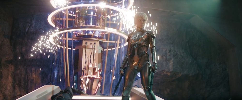 Amelia usando uma armadura metálica e atrás dela tem uma máquina soltando faíscas.