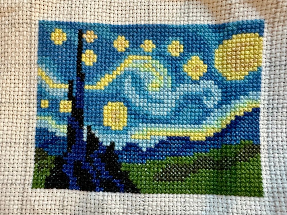 This is a cross-stitch representation of Vincent van Gogh’s “The Starry Night” in a compact, stylized format.

Key details include:
	•	Bright yellow stars and a moon against a swirling blue night sky, capturing the iconic motion of the original painting.
	•	A dark cypress tree extending vertically on the left, standing out against the vivid background.
	•	Rolling green hills and a hint of buildings or landscape at the bottom.

The piece showcases a simplified yet recognizable version of the masterpiece, with vibrant colors and detailed stitching to bring it to life.