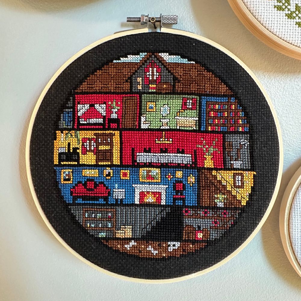 Framed cross stitch on black aida cloth in a wood hoop. Stitching is of a creepy Victorian style doll house, as seen from the front looking into each room.