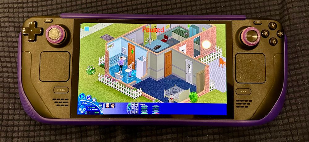 This image shows a Steam Deck handheld gaming device with purple joystick covers and a matching protective case. The device’s screen is displaying gameplay from The Sims 1, a life simulation game. The game is paused, as indicated by the red “Paused” text at the top of the screen. The view shows a house interior with various rooms, including a bathroom, kitchen, and bedroom, along with Sims characters performing actions. The bottom interface displays the classic Sims needs meters and control buttons. The device is placed on a dark textured surface.