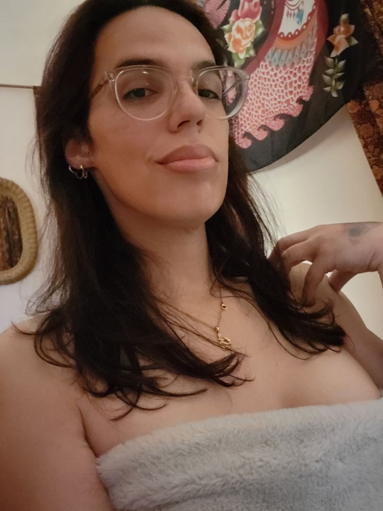 selfie of me in bed, wrapped in my blanket. im smirking. my black hair hangs down onto my shoulders. litte gold earring chains and a gold necklace are the accents