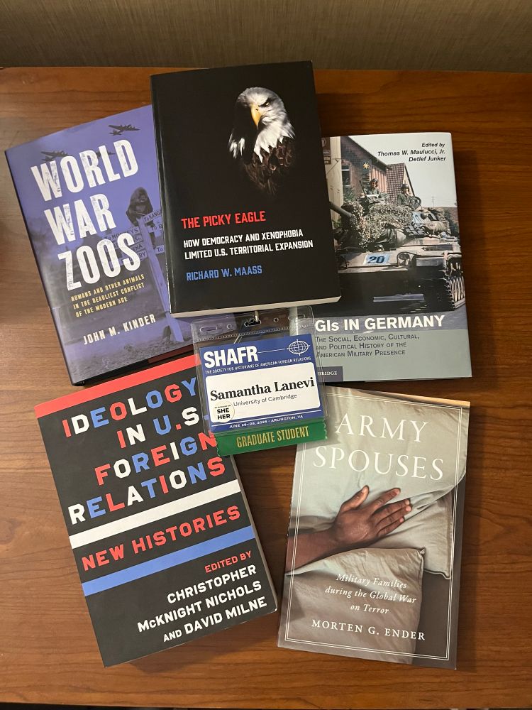 4 books related to military history, foreign relations, and 1 book related to zoos during wartime with a conference badge displayed in the middle on a wooden table