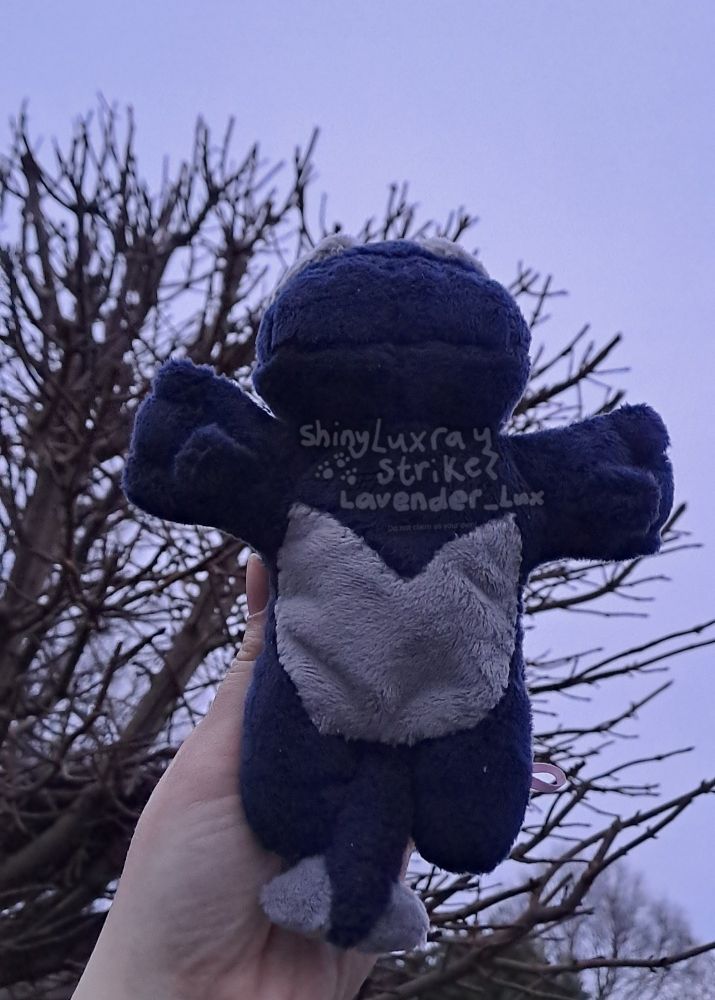 A shadow Lugia (or dark Lugia, a very dark blue version of Lugia with grey accents) kuttari (beanie) held up against a backdrop of dark tree branches and a gloomy morning sky. This picture is taken from its bottom angle.