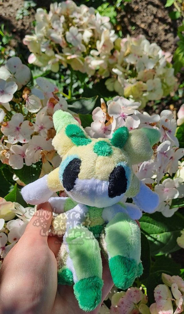 My hand holding a custom shiny Lurantis plush in chibi/pokedoll style in front of some white flowers.
