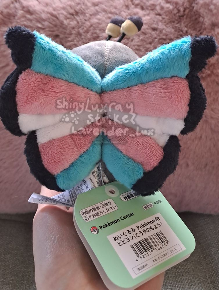 A Vivillon plush with custom wings made by me in the trans pride flag colors, showing the backside. They are held up against a backdrop of a green couch with a pink fluffy pillow