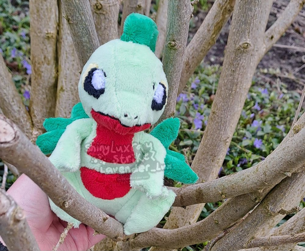A chibi Grovyle plush I made for an arttrade, sitting in a tree with some purple flowers in the background. Base pattern is by cholyknight w edits by me!