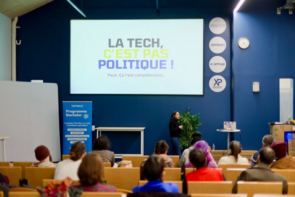 Marcy dans l’Amphi d’Epitech Lyon en train de donner sa conférence au sujet du techno autoritarisme dans le design persuasif. On peut voir l’audience et affichée en grand l’inscription suivante : « La tech c’est pas politique ! » « faux, ça l’est complètement »