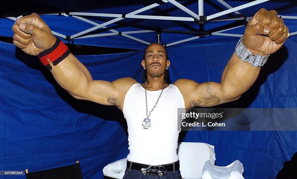 Ludacris wearing a white tank top and necklace with his arms raised and outstretched. He is wearing a prosthetic to make his forearms preposterously large