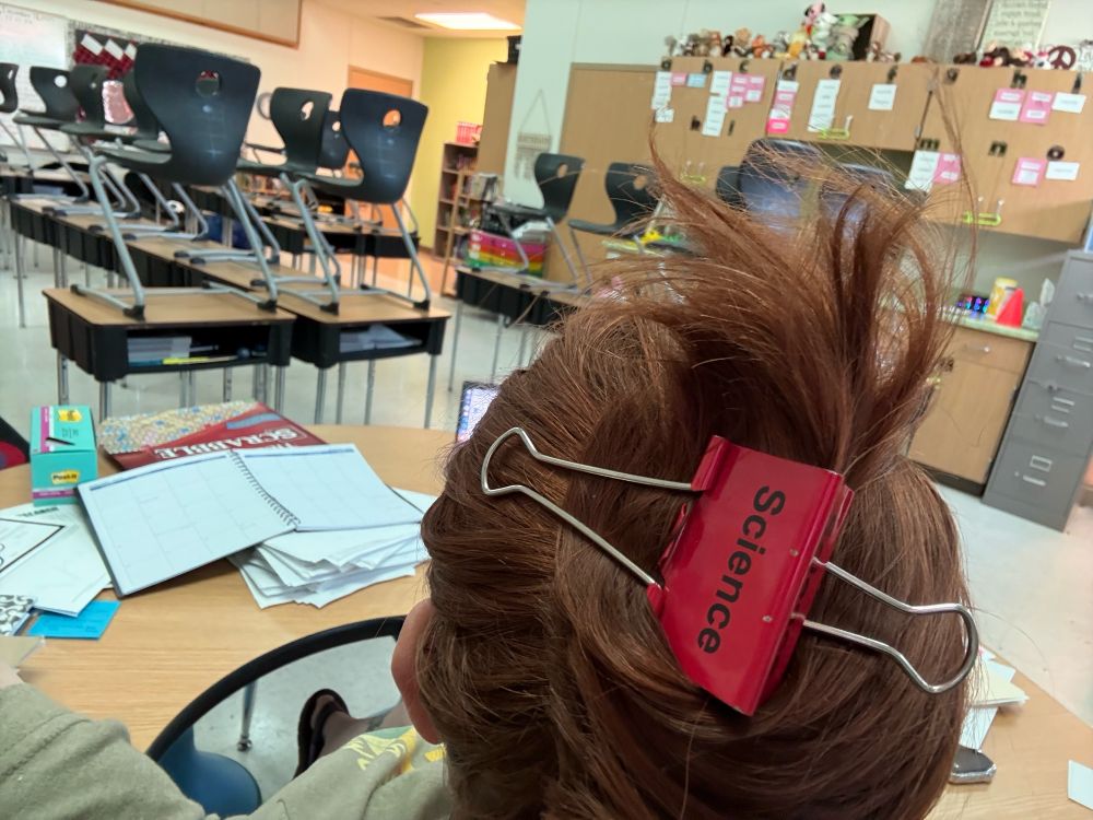 Red hair pulled up with a red binder clip that says science on it. In a classroom.