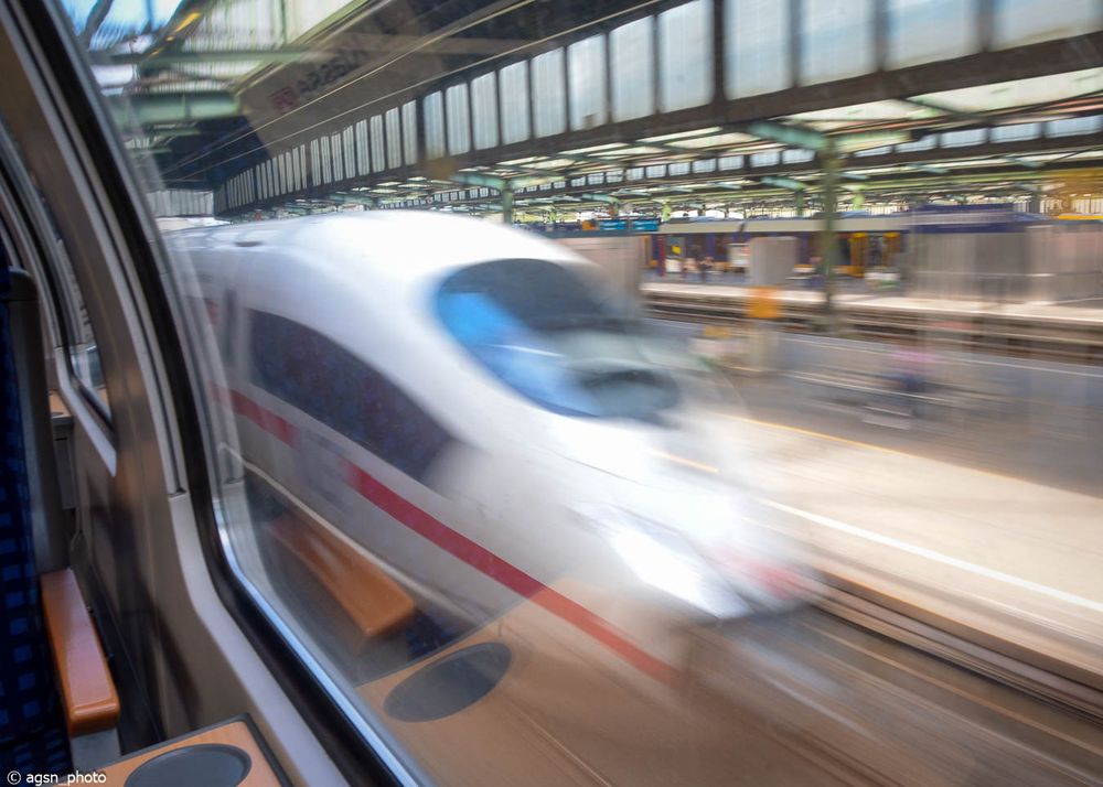 Blick aus dem Fenster eines in einem Bahnhof stehenden Zuges. Auf dem Nebengleis verlässt ein ICE mit hoher Geschwindigkeit den Bahnhof. / View from the train window. On the adjacent track, an ICE express train is leaving the station at high speed. 