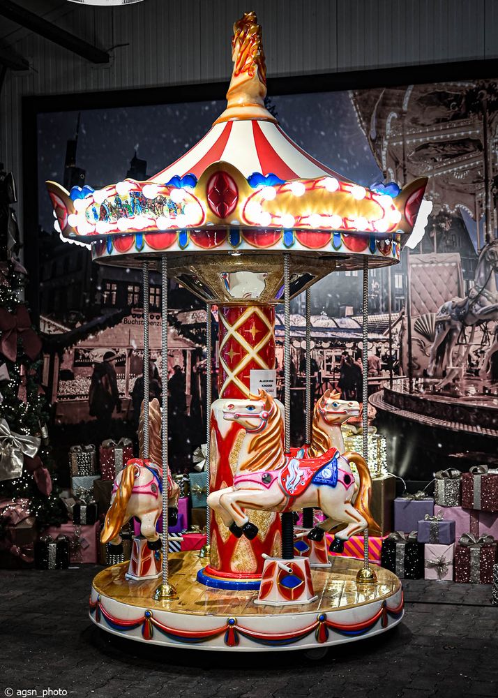 Kleines, buntes Karussell mit drei Pferden und rot-weiß gestreiftem Dach vor einem schwarz-weißen Hintergrund mit Weihnachtsmotiven. /
Small colorful carousel with three horses and a red-and-white striped roof in front of a black-and-white Christmas-themed background.
