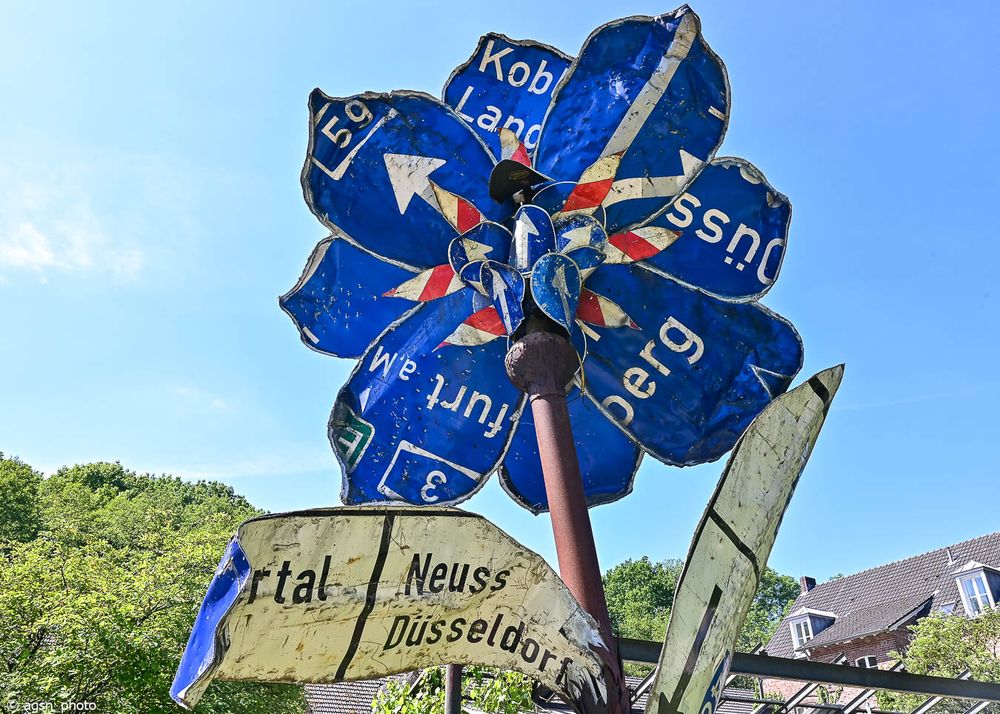 Alte blaue Autobahnschilder wurden zu einer großen Blume zusammengeschweißt, die in den blauen Himmel ragt. / Old blue motorway signs were welded together to form a large flower that rises into the blue sky. 