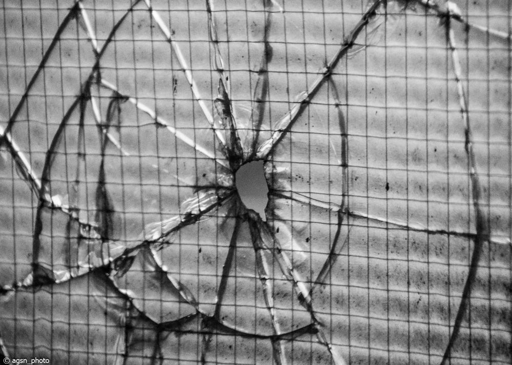 Nahaufnahme eines zerbrochenen Fensters mit einem Loch in der Mitte und sichtbaren Rissen im Glas. /
Close-up of a broken window with a hole in the center and visible cracks in the glass.
