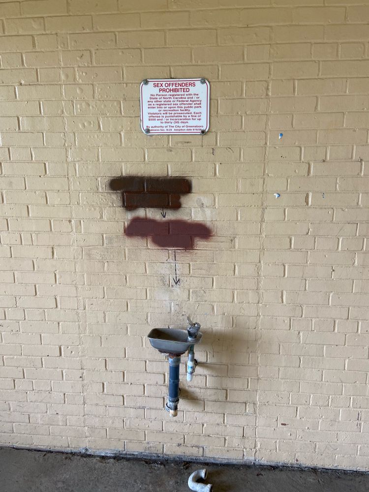 A wall in a public park with a broken water fountain and a sign saying “sex offenders prohibited”