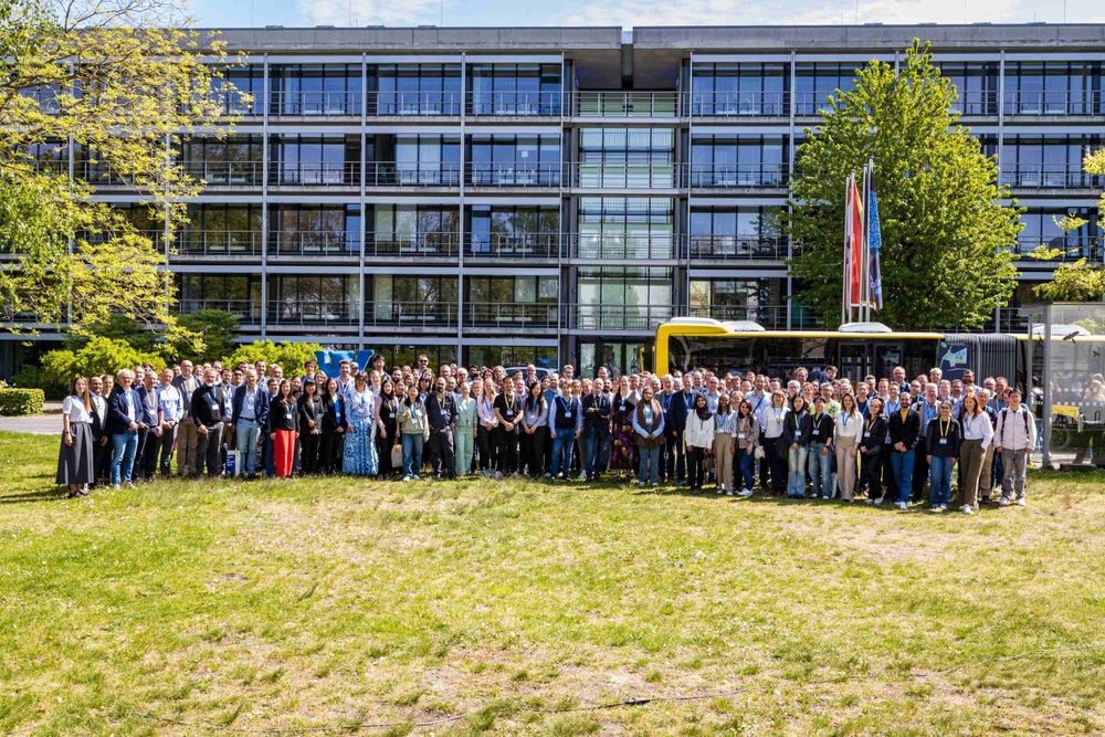 A group portrait outdoors: The image shows a large group of people standing on a grassy lawn in front of a modern glass-and-concrete building with multiple balconies. A yellow bus is parked behind the group, partially visible, along with flagpoles displaying several flags. Most individuals are facing the camera, wearing name badges and conference lanyards.