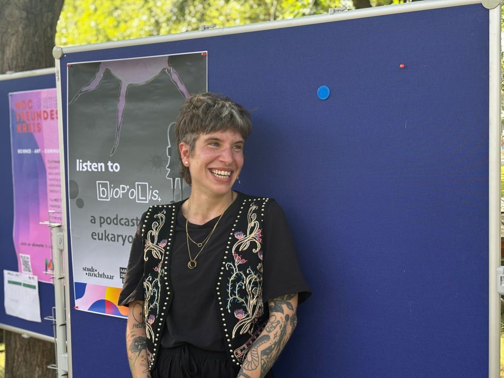 Person smiling in front of a “biopolis” podcast poster at an outdoor event.