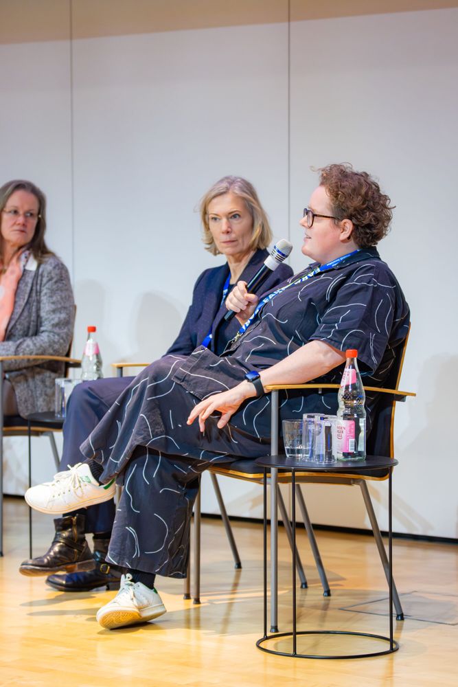 The image provides a close-up view of three of the panelists. One speaker is holding a microphone while the others listen. Bottles of water and glasses are placed on side tables next to them.