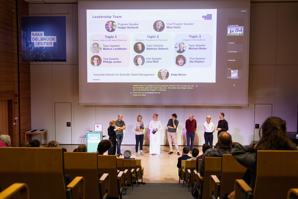 Eight people standing in front of an audience inside a lecture hall. There is a presentation above them and a banner hanging from the ceiling showing the logo of the Max Delbrück Center.
