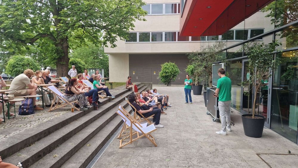Outdoor presentation with seated audience listening to two speakers near a building.