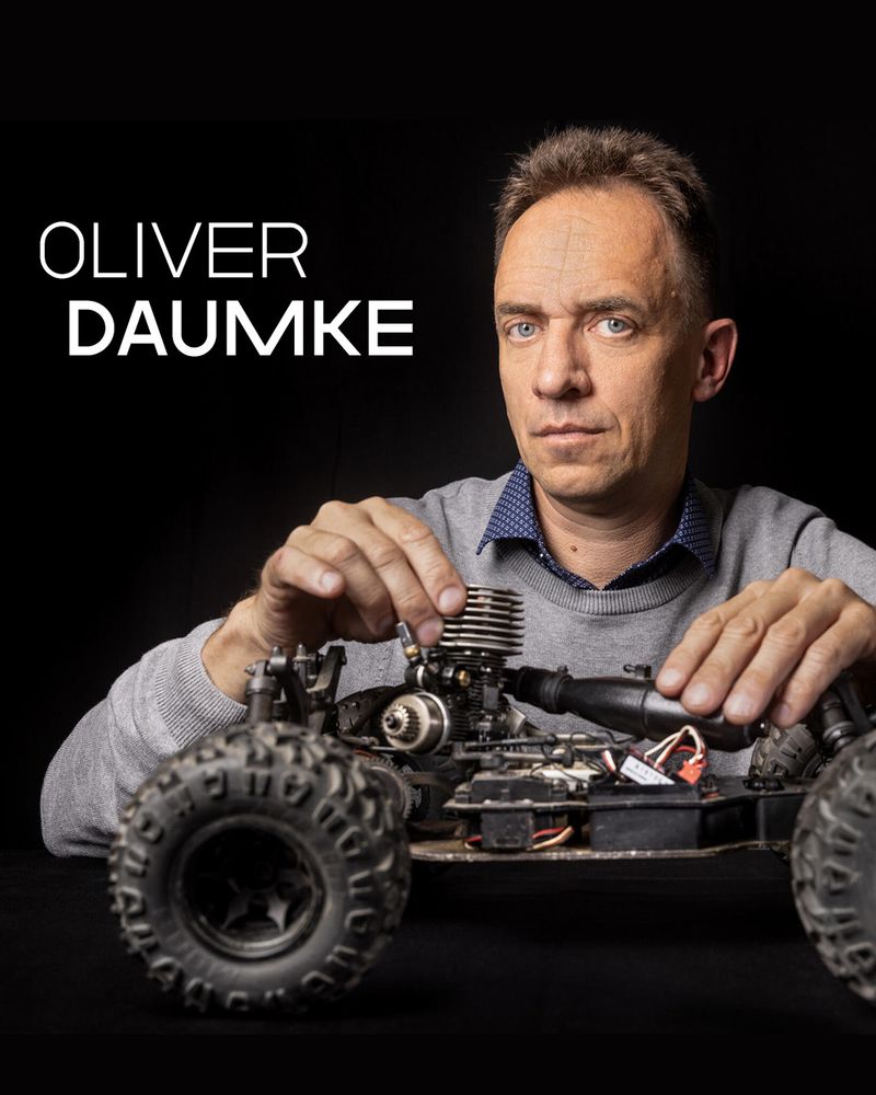 Portrait photo with text overlay: The image shows a man in a grey sweater working on a large remote-controlled car, positioned on a black table with a dark background. He looks directly at the camera while adjusting the car’s engine. In the top left corner, white sans-serif text reads “OLIVER DAUMKE,” with “DAUMKE” in bold.