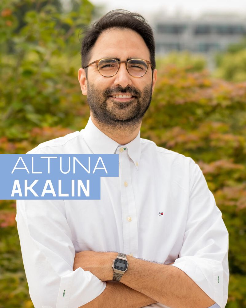 Portrait photo with text overlay: The image shows a man in a white button-down shirt standing outdoors with arms crossed, facing the camera and smiling slightly. The background is filled with greenery and blurred buildings. Overlaid text in white sans-serif font appears in the lower left corner, reading “ALTUNA AKALIN,” with “AKALIN” in bold uppercase, all inside two light blue text boxes.