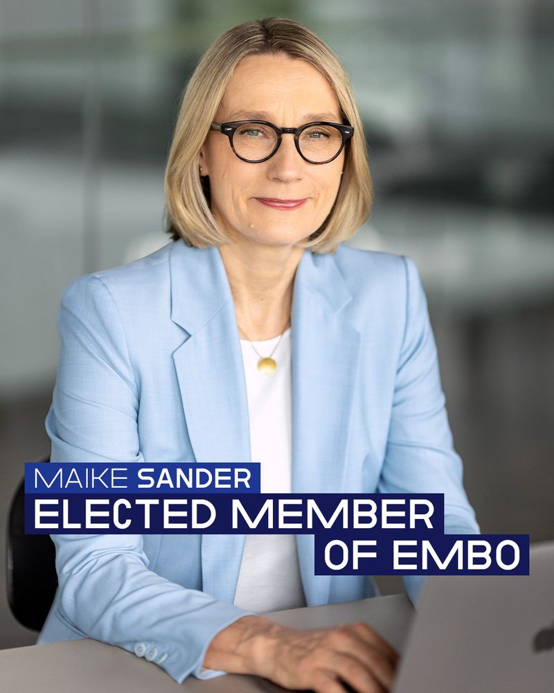 Portrait photo with text overlay: The image shows a woman [Maike Sander] in a light blue blazer and glasses seated at a desk with a blurred office background. She is looking at the camera with a neutral expression, hands on a laptop.

Overlaid text in bold white sans-serif font appears across the lower half, set against dark blue rectangles. It reads, “MAIKE SANDER ELECTED MEMBER OF EMBO,” with “MAIKE SANDER” in smaller uppercase and the rest in larger all-caps.