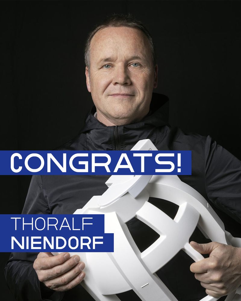 Portrait photo with text overlay: The image shows a man in a black jacket standing against a dark background, holding a large white sculptural object. He is looking directly at the camera with a neutral expression. Bold white sans-serif text appears in blue text boxes across the image, reading “CONGRATS!” in the center and “THORALF NIENDORF” in the lower left, with “NIENDORF” in bold uppercase.