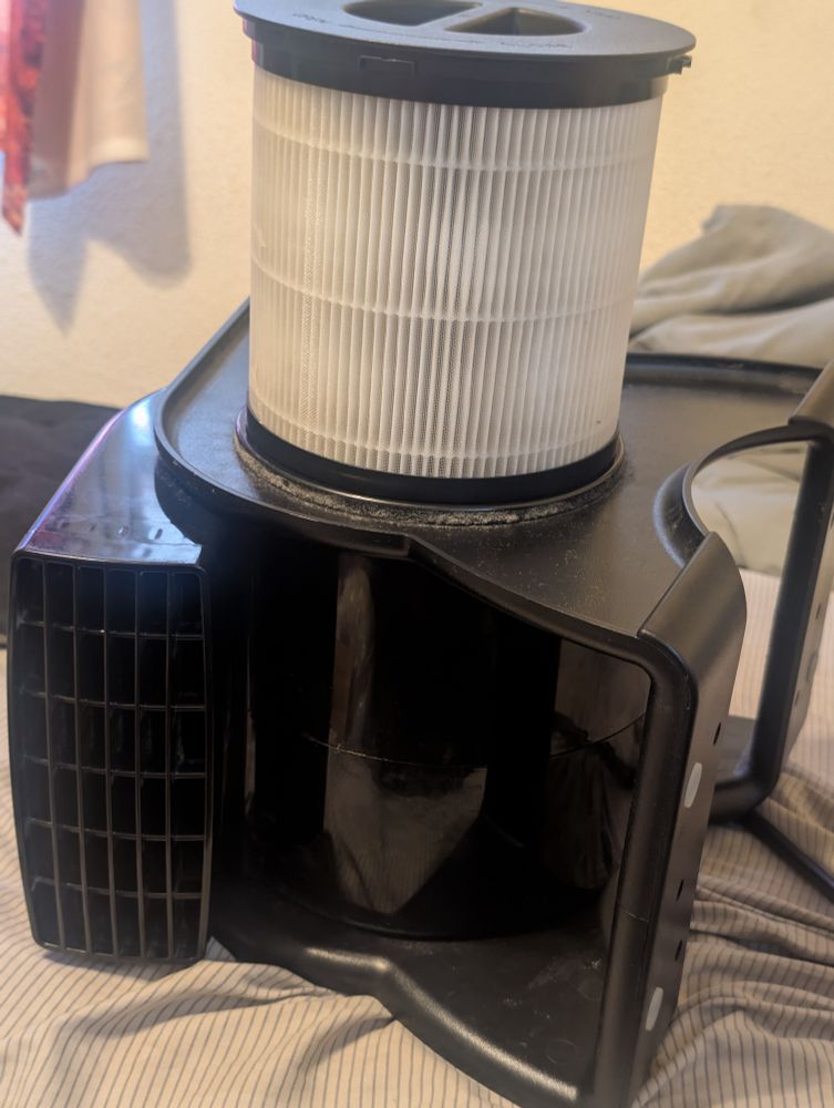 Photo of a blower style fan sitting on its side on a bed with a round HEPA filter sitting on top, and a cap on top of the filter. The filter and cap came from a broken air purifier. The fan is sitting on its side partly because I have no way to secure the filter (the suction helps seal it but the weight of the filter is too much for it to be held by vacuum) and partly because it pulls air from the other side, likely as a way to keep the motor cool. ... Which is probably bad to block off but the air being sucked in is bypassing the filter and reducing the purifying capabilities.
