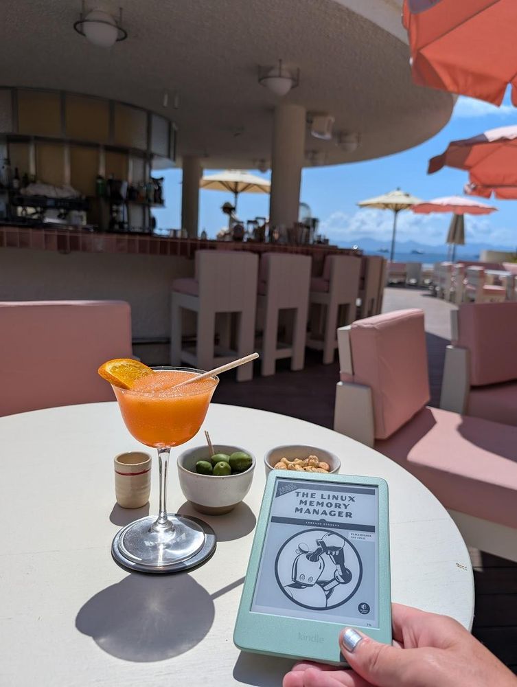 The Linux Memory Manager book displayed on a Kindle e-reader next to a cocktail on a table in a beach side bar