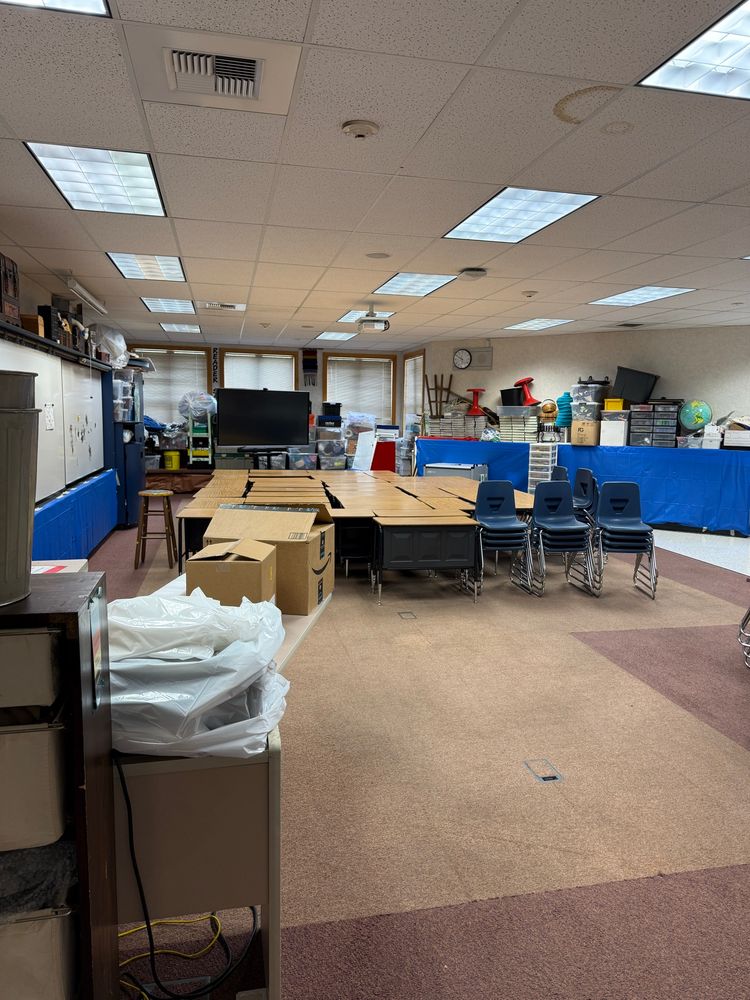 A school classroom that hasn’t been set up yet has blue plastic covering shelves, desks clustered on one side of the room and on the window bench seats are plastic tubs, bags, boxes, and tool boxes. 