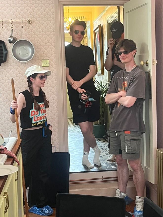 Four crew members of “A Figment of Existence” in the kitchen. Producer Lily Allmark (left) holding a broom and wearing sunglasses. PA/BTS Gareth Llewellyn in the middle, holding a camcorder, wearing sunglasses and looking at the camera. On the front right spark Noël Hoffmanns is wearing sunglasses and has his arms folded as he looks at the camera. Behind him, the other spark Oliver Williams is hiding under his cap. They are surrounded by black flags as they are about to break a plate for the next scene.