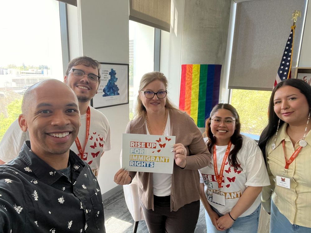 Two GCIR staff members stand with members of the California Work and Family Coalition at the California Immigrant Day of Action 2025 event. One of the coalition members is holding a colorful sign of red, green, yellow, and blue text that reads: "Rise Up For Immigrant Rights." The sign has red butterflies in the upper right corner.