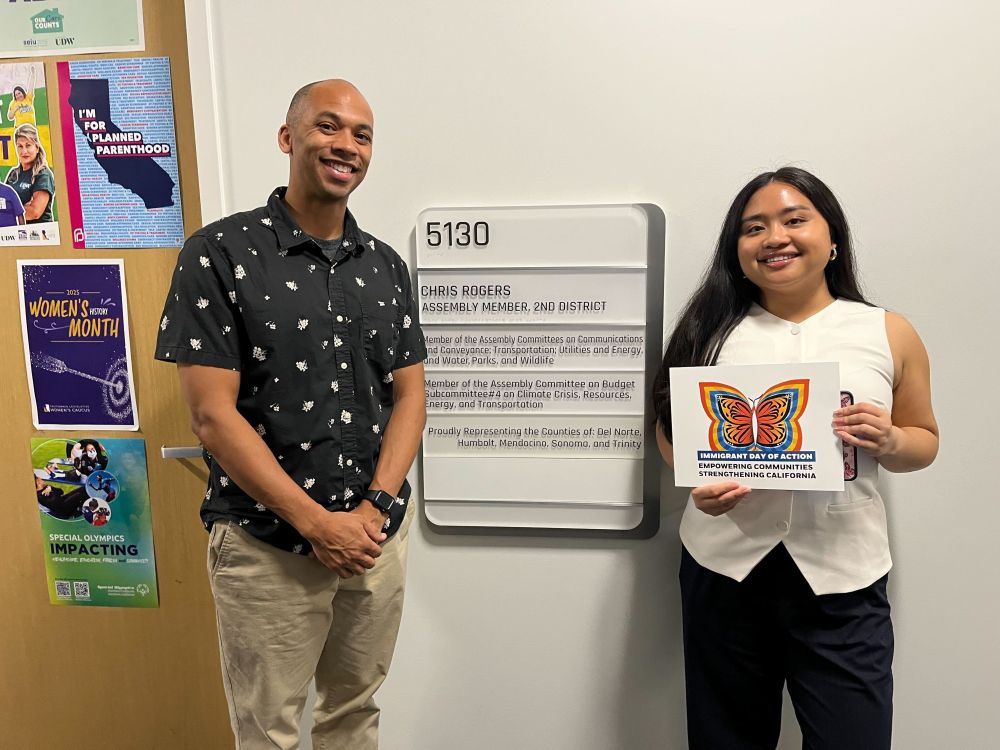  A GCIR staff member stands with a staff member from the office of California Assembly Member Chris Rogers at the California Immigrant Day of Action 2025 event. The assemblymember's staff member is holding a sign with a yellow, blue and orange butterfly that reads, "Immigrant Day of Action: Empowering Communities. Strengthening California."