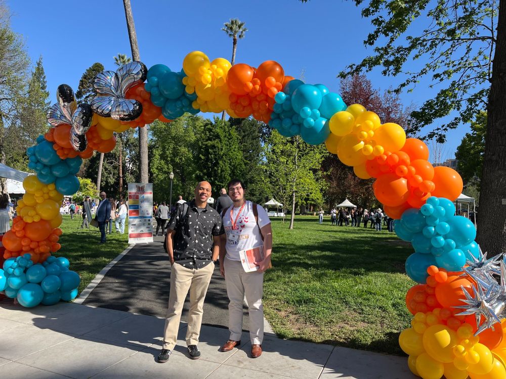Two GCIR staff members stand outside underneath a colorful arch of yellow, blue and orange balloons at the California Immigrant Day of Action 2025 event.