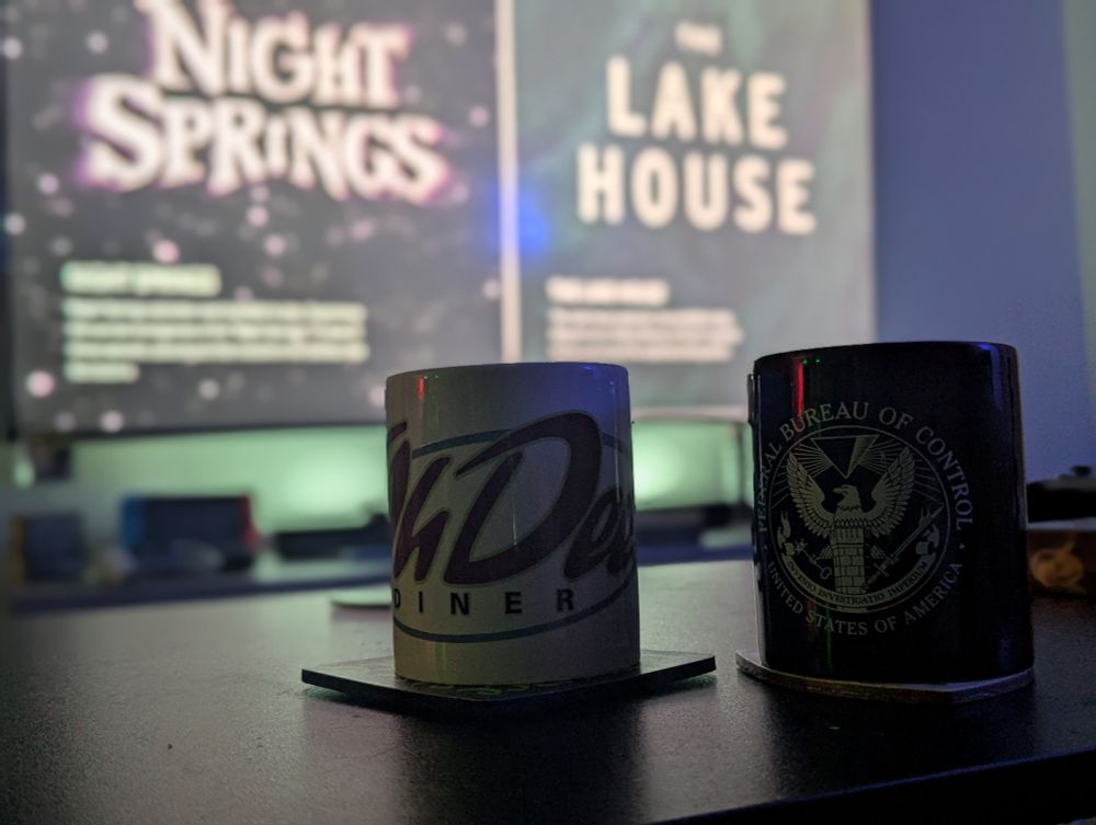 two coffee mugs in front of a television screen. 
 
One with the "Oh Deer Diner"  logo from the video game Alan Wake. 

The second had the badge of the "Federal Bureau of Control" from the video game CONTROL. 

On the television screen is the Alan Wake 2 DLC option screen for "Night Springs" and "The Lake House" 