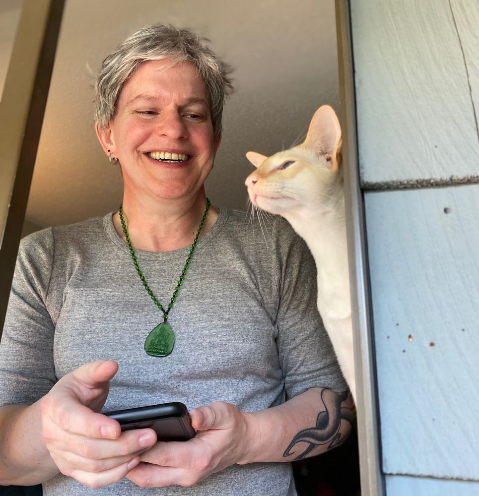 Middle-aged woman with short grey hair laugh-smiling at a thin white cat poking his nose out the window.