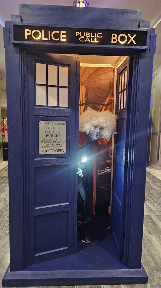 Cosplay of Strive, a blue alien with white hair, from the music video for Starlight Brigade, peekong out of the Tardis from Dr. Who.
