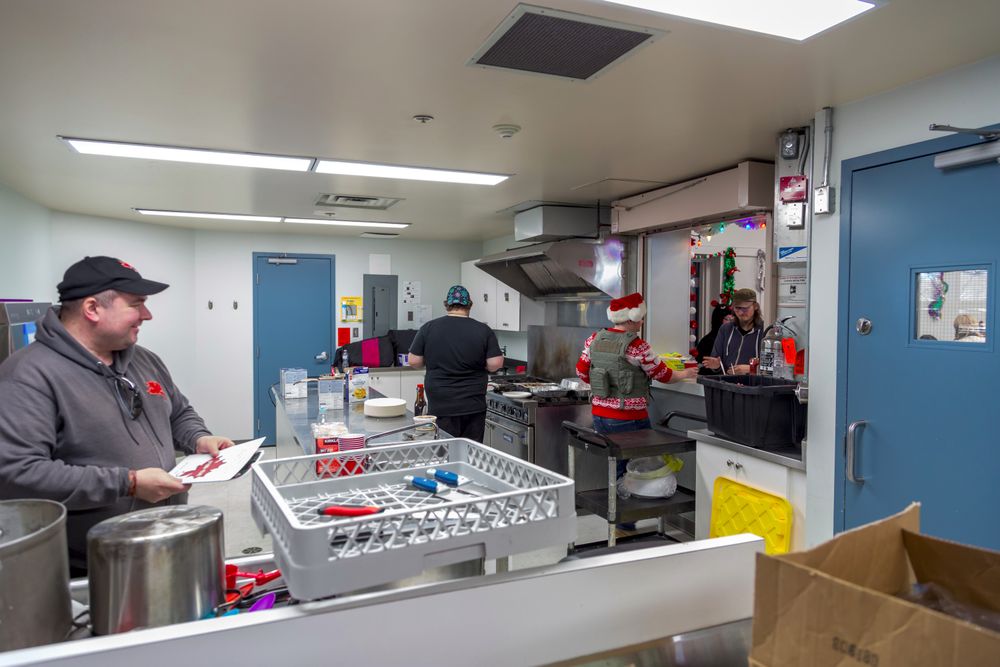 Three volunteers working in the kitchen at the 2024 Fur-Eh! Christmas Party. One is holding a small sign and smiling, one is at the stove with their back to the photographer, and one is at the counter window serving a drink to a guest.