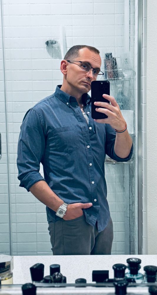 dumb guy taking a sneaky lil mirror selfie before heading out with friends. he's wearing glasses, a light blue button-down, grey pants, and a silver watch.