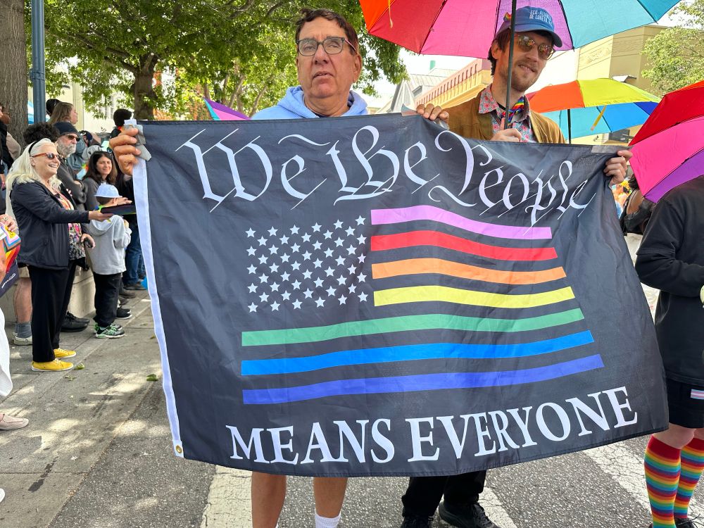 Street scene parade photo of a banner that reads, “We the people MEANS EVERYONE” that borders a U.S. flag with stripes colored by pride colors. 
