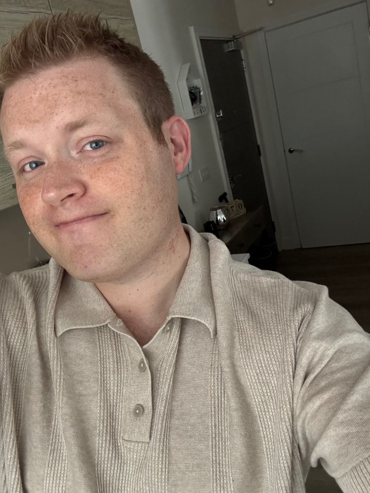 A young, blue-eyed man wearing a cream top staring and smiling directly at the camera. 