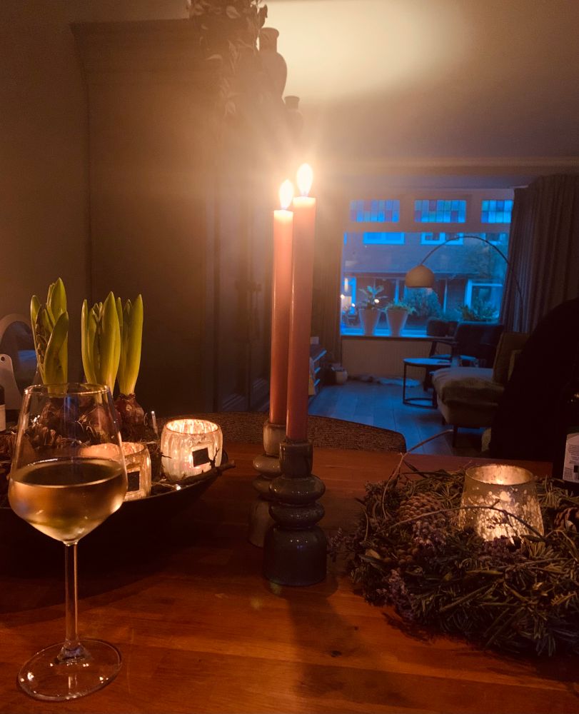 Foto van onze eetkamertafel met een glas witte wijn, 2 brandende kaarsen en een schaal met 5 bollen hyacinten, mos en dennenappels en 2 glaasjes met waxinelichtjes 